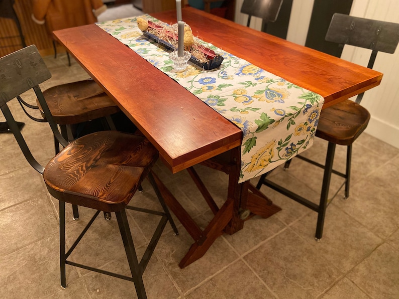Op de afbeelding: Een houten eettafel met een bloemige tafelrunner en vier zwarte metalen stoelen met houten zittingen. De tafel heeft een rustieke, landelijke stijl.
