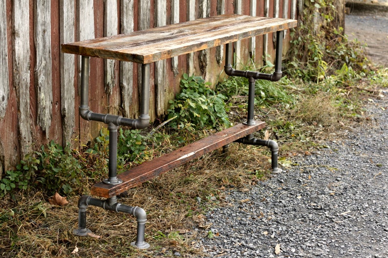 May include: A rustic wooden table with a black metal pipe frame. The table has a distressed wood top and a lower shelf. The table is standing on a gravel path.