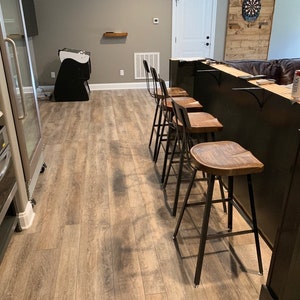 Op de afbeelding: Een zwarte bar met een houten blad en vier zwarte metalen barstoelen met houten zittingen. De bar bevindt zich in een kelder met een houten wand en een koelkast op de achtergrond.
