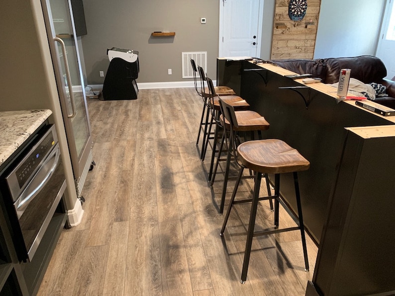 Op de afbeelding: Een zwarte bar met een houten blad en vier zwarte metalen barstoelen met houten zittingen. De bar bevindt zich in een kelder met een houten wand en een koelkast op de achtergrond.