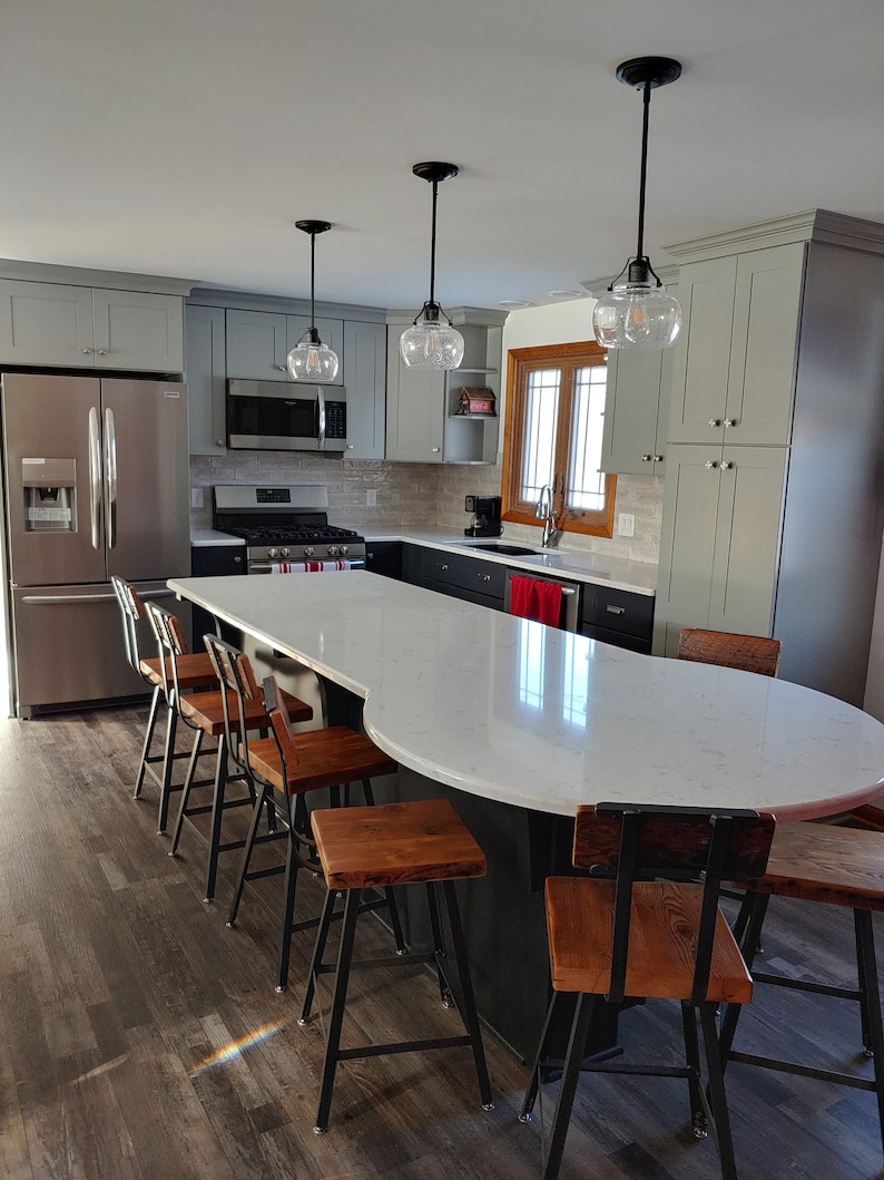 May include: A modern kitchen with gray cabinets, stainless steel appliances, and a large white quartz island with a curved edge. The island has a black base and features several wooden bar stools with black metal frames.