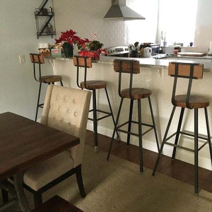 May include: Four black metal bar stools with wooden seats and backs. The stools are arranged in a row in front of a white counter. The counter has a white countertop and a white backsplash. There is a rug on the floor in front of the stools.