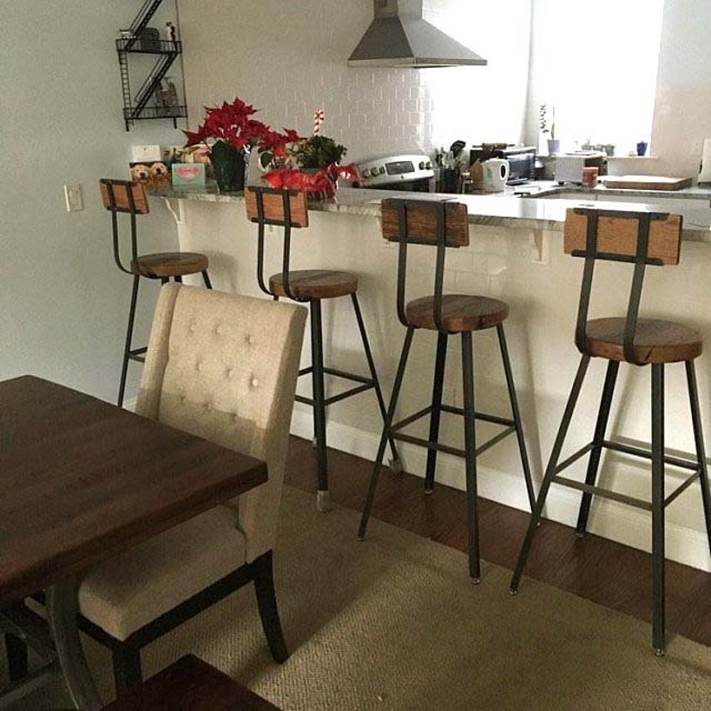 May include: Four black metal bar stools with wooden seats and backs. The stools are arranged in a row in front of a white counter. The counter has a white countertop and a white backsplash. There is a rug on the floor in front of the stools.