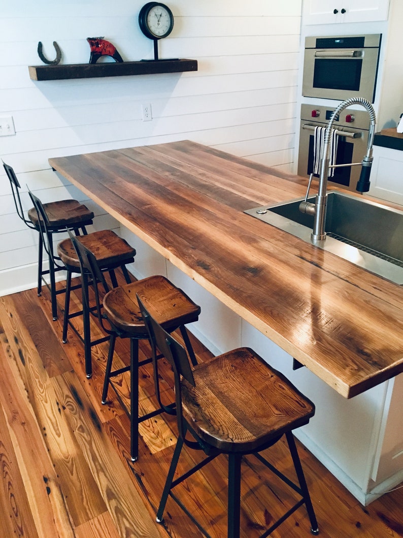 Op de afbeelding: Een keukeneiland met een werkblad van gerecycled hout en vier zwarte metalen barstoelen. Het eiland heeft een ingebouwde gootsteen en een kraan van roestvrij staal. Het werkblad is afgewerkt met een laag transparante polyurethaan.