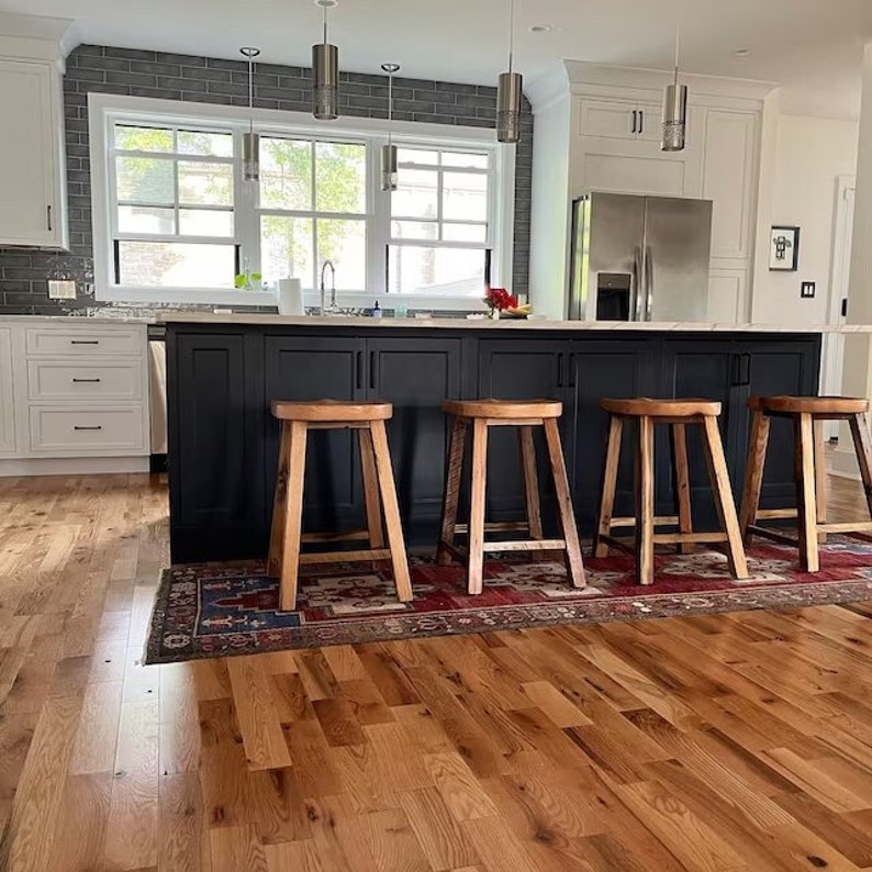 May include: A kitchen island with four wooden stools. The island is black with a light countertop. The stools are made of light-colored wood. A rug with a red and blue pattern is under the stools. The kitchen has white cabinets and a stainless steel refrigerator.