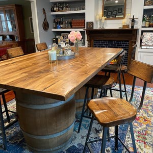 May include: A rustic wooden dining table with a light brown finish, supported by a repurposed wooden barrel base. The table is surrounded by several wooden bar stools with black metal legs. The room has a cozy, inviting atmosphere.