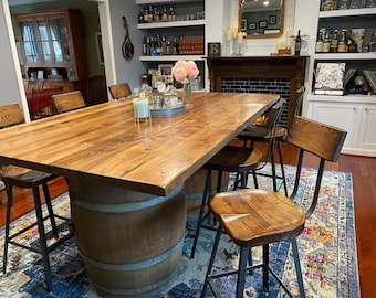 May include: A rustic wooden dining table with a light brown finish, supported by a repurposed wooden barrel base. The table is surrounded by several wooden bar stools with black metal legs. The room has a cozy, inviting atmosphere.