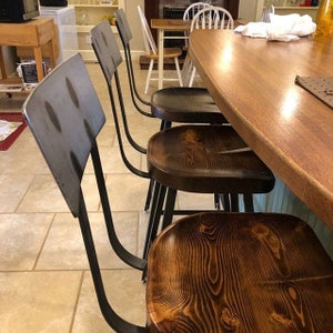 Op de afbeelding: Vier industri&euml;le barstoelen met zwarte metalen frames en houten zittingen. De zittingen zijn van donkerbruin hout met een natuurlijke houtnerf. De stoelen zijn in een rij geplaatst, met de dichtstbijzijnde stoel in focus.