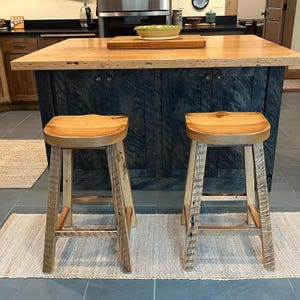 May include: Two wooden bar stools with light brown seats and light-colored, rustic legs. The stools are in front of a dark blue kitchen island with a light brown countertop. A small rug is beneath each stool.