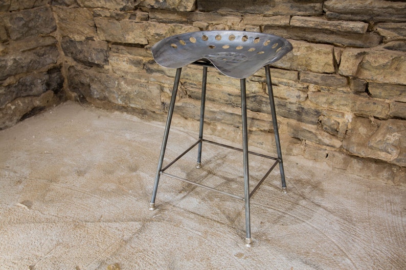 May include: A metal stool with a unique seat made from a repurposed tractor seat. The seat has a curved shape and is perforated with holes. The stool has a simple, industrial design and is made of sturdy metal.