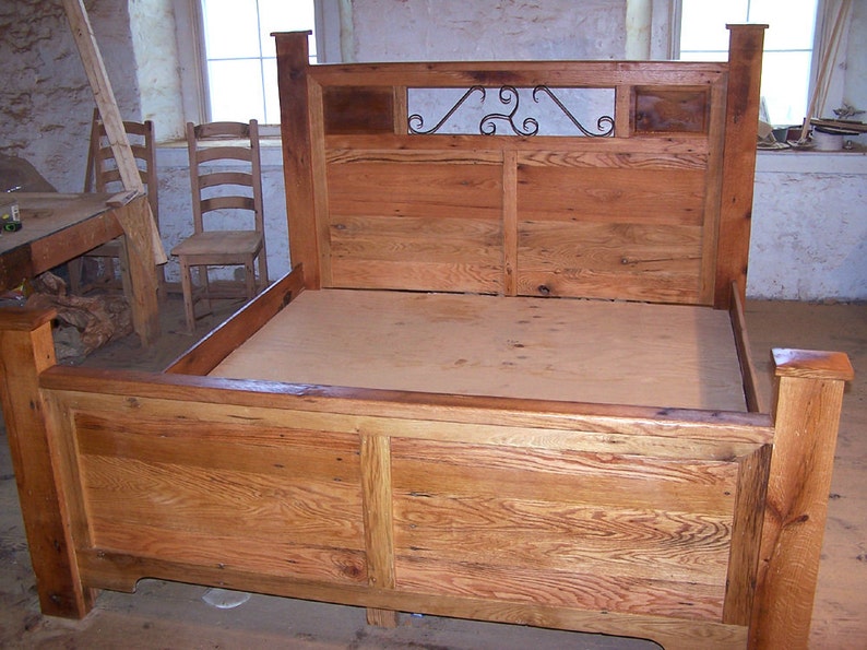 Op de afbeelding: Een rustiek houten bedframe met een hoofdbord met een decoratief metalen scroll-ontwerp. Het frame is gemaakt van gerecycled hout met een natuurlijke afwerking.