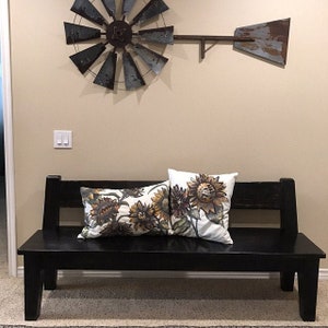 May include: A black wooden bench with two throw pillows featuring a sunflower pattern. The pillows are white with brown and yellow sunflowers. The bench is in front of a wall with a metal windmill decoration.