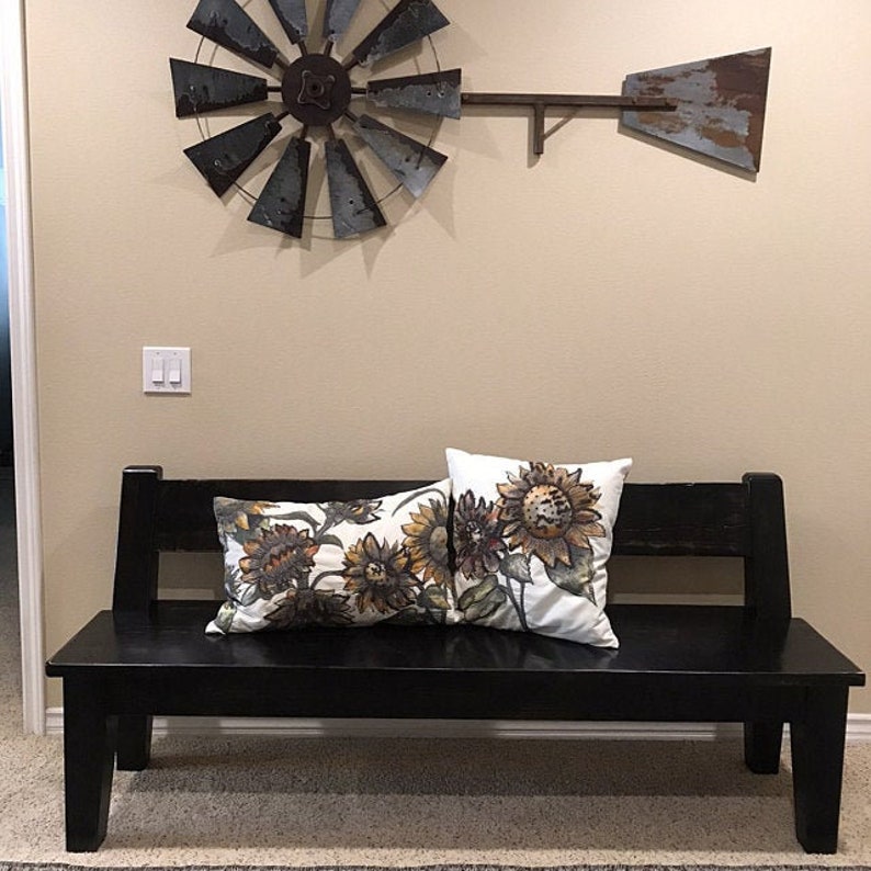 May include: A black wooden bench with two throw pillows featuring a sunflower pattern. The pillows are white with brown and yellow sunflowers. The bench is in front of a wall with a metal windmill decoration.