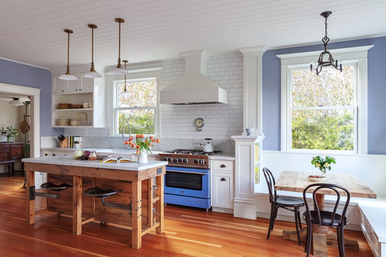 May include: A kitchen with white subway tile backsplash, a blue stove, and a wooden island with two bar stools. The kitchen has a white ceiling with wood beams, a large window with a view of trees, and a dining nook with a wooden table and chairs.