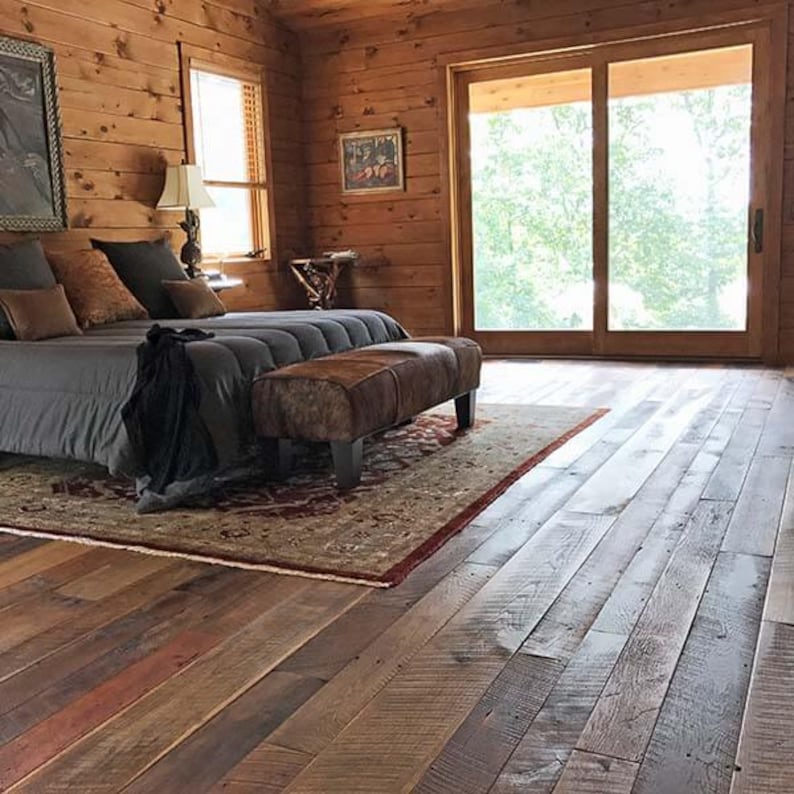 May include: A rustic bedroom with wood paneling, a bed with gray bedding, and a brown leather bench. A patterned rug sits beneath the bed, and a large window offers a view of the outdoors. The floor is made of reclaimed wood.