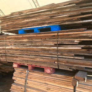 May include: A large stack of reclaimed wood planks, varying in width and length, ready for use. The wood has a weathered, aged appearance with natural color variations. The planks are stacked on blue and red pallets, secured with black straps.