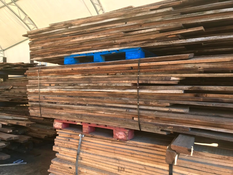 May include: A large stack of reclaimed wood planks, varying in width and length, ready for use. The wood has a weathered, aged appearance with natural color variations. The planks are stacked on blue and red pallets, secured with black straps.
