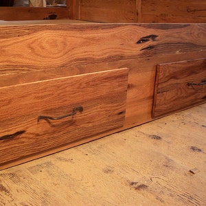 Op de afbeelding: Twee houten laden met metalen handvatten zijn bevestigd aan de onderkant van een houten bedframe. De laden zijn gemaakt van gerecycled hout met een natuurlijke afwerking.