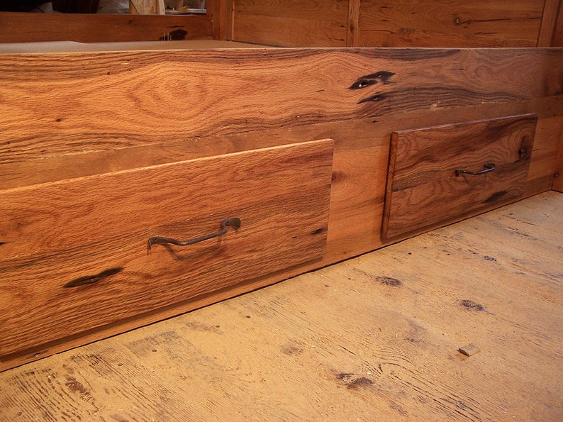 Op de afbeelding: Twee houten laden met metalen handvatten zijn bevestigd aan de onderkant van een houten bedframe. De laden zijn gemaakt van gerecycled hout met een natuurlijke afwerking.