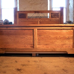 Op de afbeelding: Een houten bedframe met een hoofdbord met een decoratief metalen scroll-ontwerp. Het frame is gemaakt van gerecycled hout met een natuurlijke afwerking.