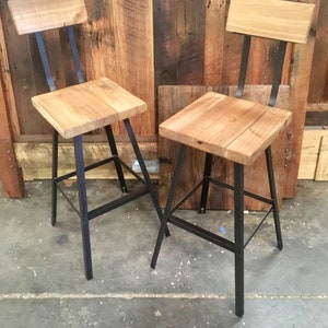 May include: Two wooden and metal bar stools with black metal frames and a natural wood finish. The stools have a simple design with a rectangular seat and a back rest.