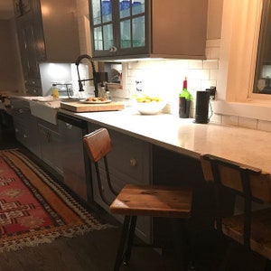 May include: A kitchen counter with a white countertop and gray cabinets. There are two wooden bar stools with black metal frames. A sink, a faucet, and a cutting board are also visible.