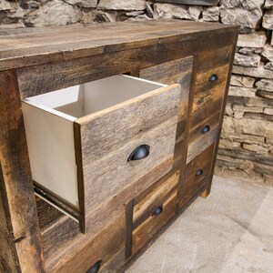 May include: A rustic wooden dresser with multiple drawers, one of which is open, revealing a white interior. The dresser has a distressed wood finish and black metal drawer pulls.