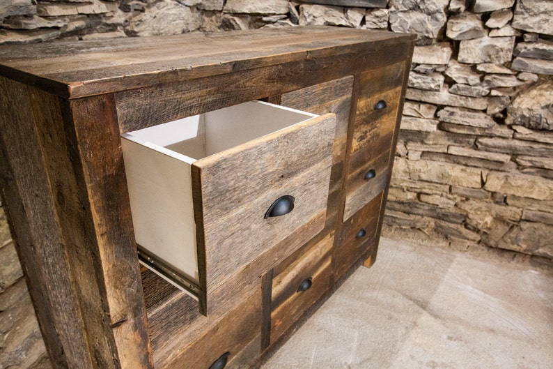 May include: A rustic wooden dresser with multiple drawers, one of which is open, revealing a white interior. The dresser has a distressed wood finish and black metal drawer pulls.