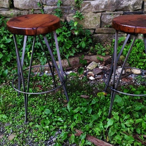 May include: Two wooden and metal bar stools with a rustic wood seat and a black metal frame. The stools are set against a stone wall and green foliage.