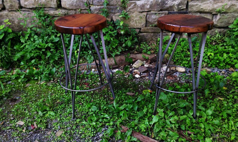 May include: Two wooden and metal bar stools with a rustic wood seat and a black metal frame. The stools are set against a stone wall and green foliage.
