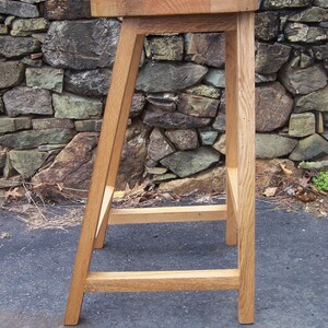 May include: A handcrafted wooden bar stool with a square seat and angled legs. The stool is made of light-colored wood and has a simple, minimalist design. The stool's height is approximately 30 inches.