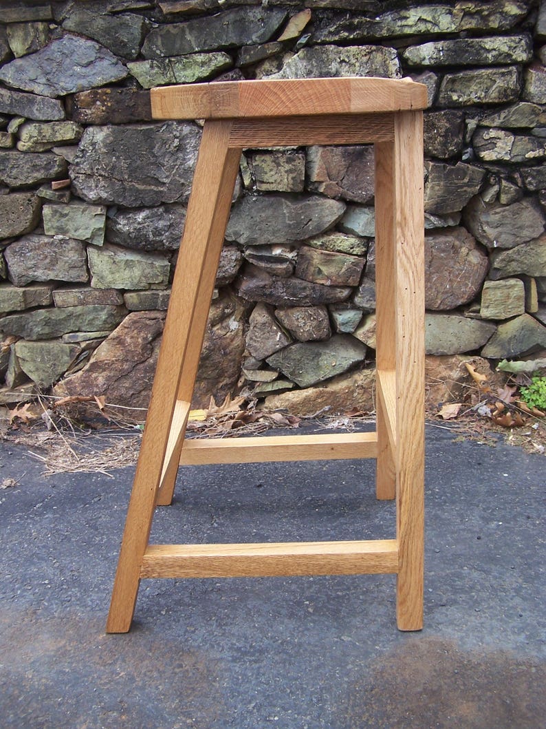 May include: A handcrafted wooden bar stool with a square seat and angled legs. The stool is made of light-colored wood and has a simple, minimalist design. The stool's height is approximately 30 inches.