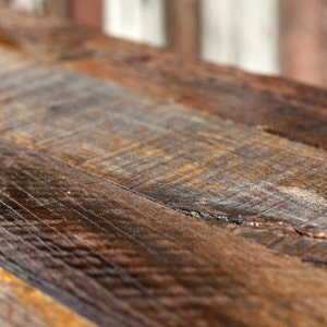 May include: Close-up of a rustic wooden table top with a natural wood grain pattern. The wood is a mix of brown and dark brown shades, with a few knots and imperfections.
