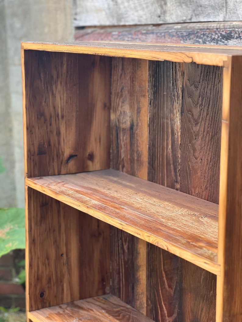 May include: A rustic, wooden bookshelf with two shelves. The wood has a natural, warm brown tone with visible grain and knots, giving it a reclaimed or vintage appearance. The shelves are sturdy and ready for display.