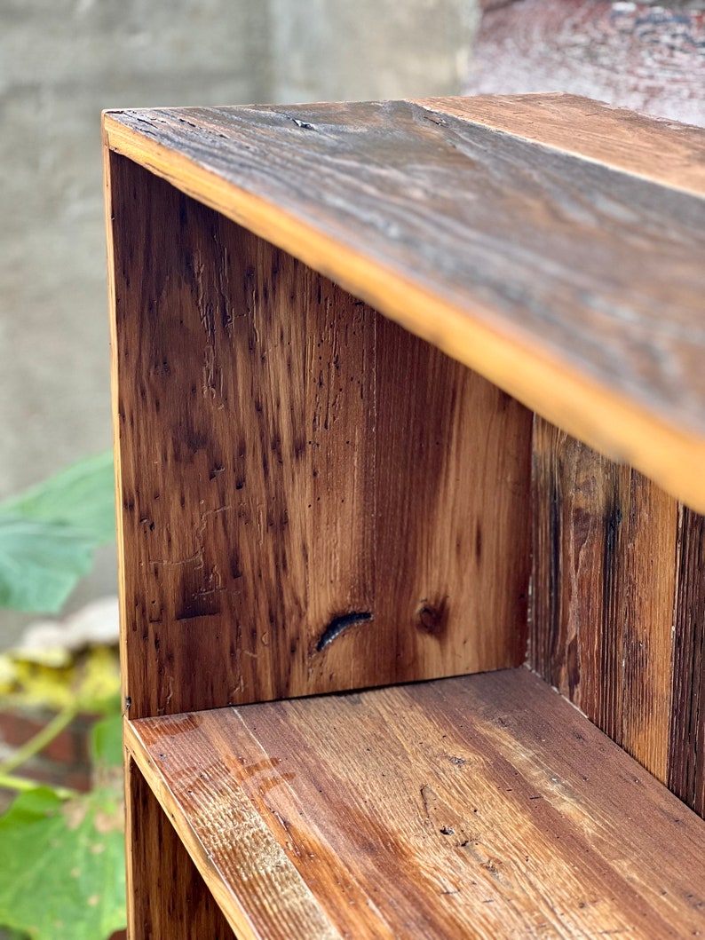 May include: Close-up of a rustic wooden shelf unit. The unit is constructed from reclaimed wood, showcasing a mix of light and dark brown tones with visible wood grain and knots. The design features a simple, open-shelf structure, perfect for displaying items.