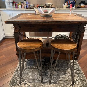 May include: Two rustic wooden bar stools with metal legs and a distressed finish. The stools are placed on a patterned rug in front of a wooden kitchen island.