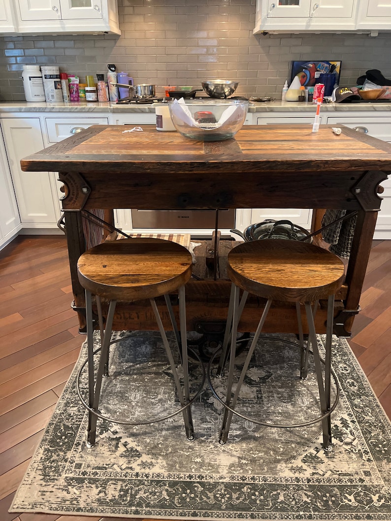 May include: Two rustic wooden bar stools with metal legs and a distressed finish. The stools are placed on a patterned rug in front of a wooden kitchen island.