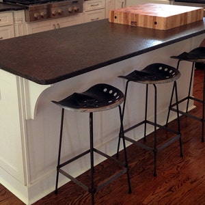 May include: A white kitchen island with a dark brown granite countertop and a wooden cutting board. Three black metal stools with a unique seat design are positioned around the island.