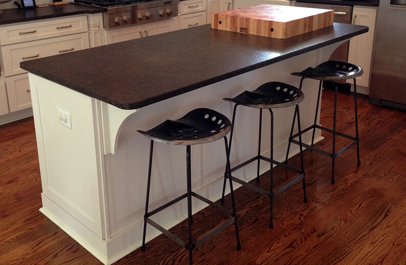 May include: A white kitchen island with a dark brown granite countertop and a wooden cutting board. Three black metal stools with a unique seat design are positioned around the island.