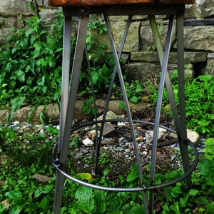 May include: A rustic wooden stool with a dark brown finish and a metal frame. The stool has a circular seat and a tripod base. The stool is sitting on a bed of green grass.