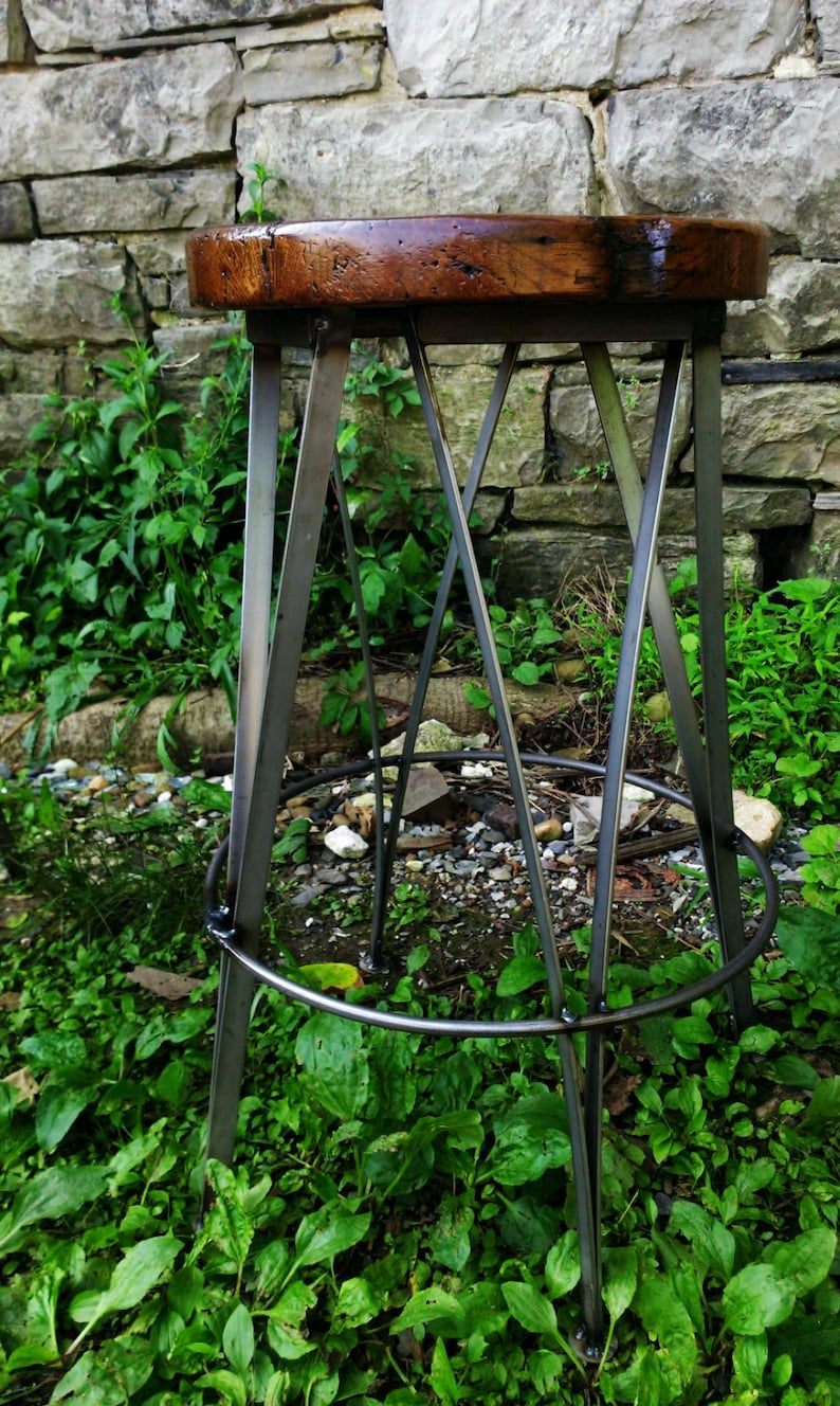 May include: A rustic wooden stool with a dark brown finish and a metal frame. The stool has a circular seat and a tripod base. The stool is sitting on a bed of green grass.