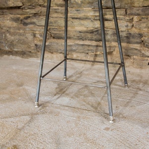May include: A metal bar stool with a triangular base and a square footrest. The stool has a silver finish and is sitting on a concrete floor.