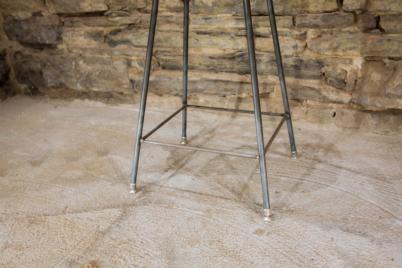 May include: A metal bar stool with a triangular base and a square footrest. The stool has a silver finish and is sitting on a concrete floor.