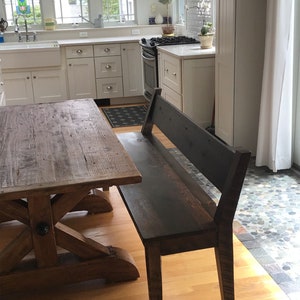 May include: A rustic wooden table with an X-shaped base and a dark wood bench with a back rest. The table and bench are in a kitchen with white cabinets and a tiled floor.