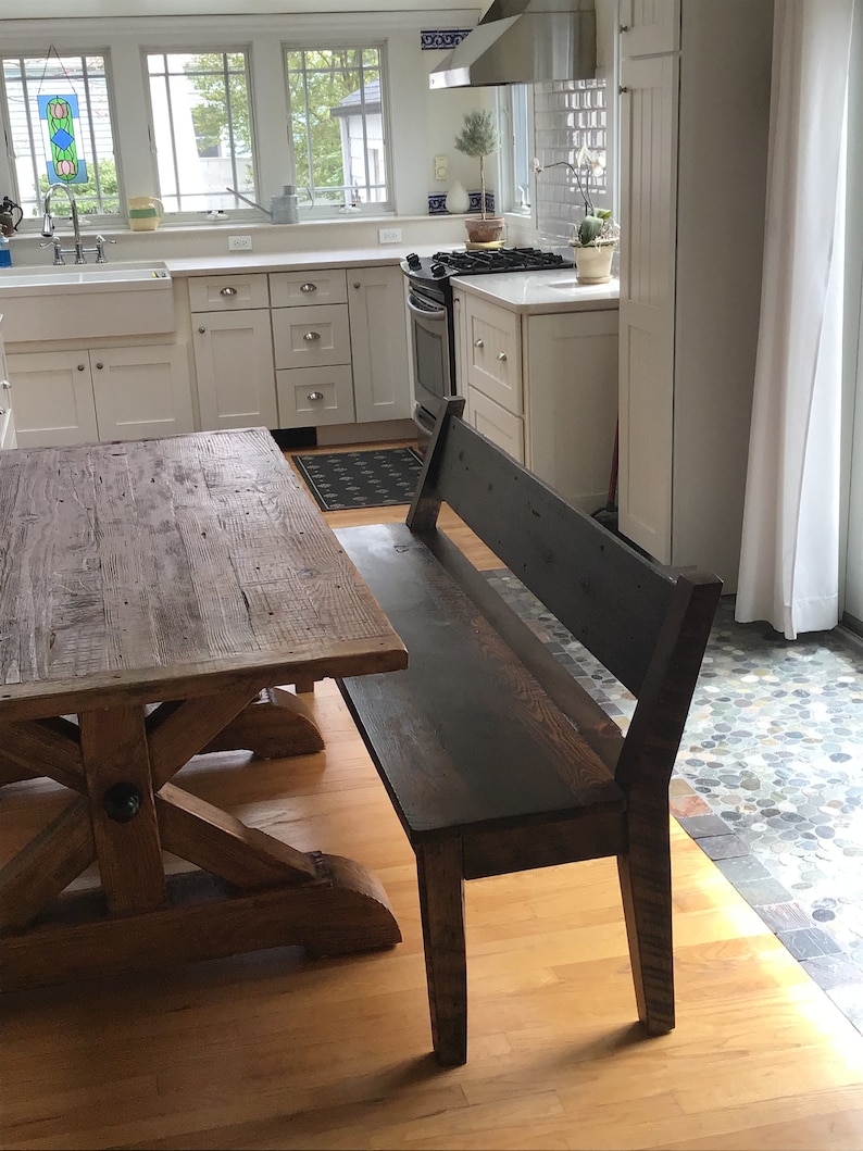 May include: A rustic wooden table with an X-shaped base and a dark wood bench with a back rest. The table and bench are in a kitchen with white cabinets and a tiled floor.