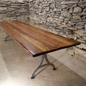 May include: A long, rustic wooden table with a natural finish. The table has a thick, wide top and is supported by four metal legs.