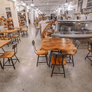 May include: Interior view of a cafe or market featuring wooden tables and chairs. The tables have a natural wood finish and are paired with black metal chairs. The floor is concrete, and the ceiling has exposed beams and hanging lights.