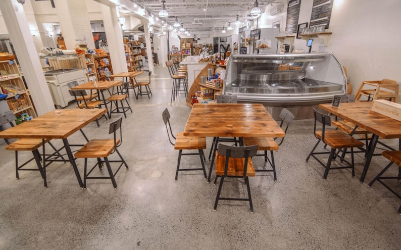 May include: Interior view of a cafe or market featuring wooden tables and chairs. The tables have a natural wood finish and are paired with black metal chairs. The floor is concrete, and the ceiling has exposed beams and hanging lights.