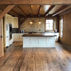 May include: Interior view of a rustic kitchen with wooden floors, walls, and ceiling beams. The kitchen features white cabinets, a central island, and a brick backsplash. Two pendant lights hang above the island, and windows provide natural light.