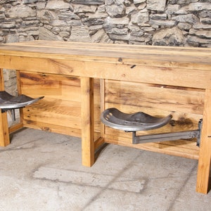 May include: A rustic wooden bar table with two metal tractor seat stools attached to the underside. The table has a natural wood finish and is made from reclaimed wood. The stools are black metal and have a simple design.